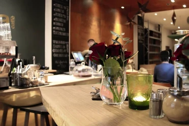 moxxa cafe die rösterei Ein eleganter Café-Innenraum mit Holztheken, auf denen rote Rosen in Glasvasen stehen, während Kerzenlicht eine gemütliche Atmosphäre erzeugt. Im Hintergrund eine Kaffeetheke.