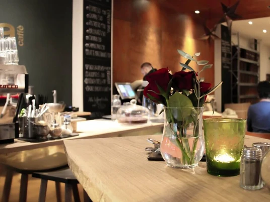 moxxa cafe die rösterei Ein eleganter Café-Innenraum mit Holztheken, auf denen rote Rosen in Glasvasen stehen, während Kerzenlicht eine gemütliche Atmosphäre erzeugt. Im Hintergrund eine Kaffeetheke.An elegant café interior with wooden counters adorned with red roses in glass vases, while candlelight creates a cosy atmosphere. A coffee bar can be seen in the background.