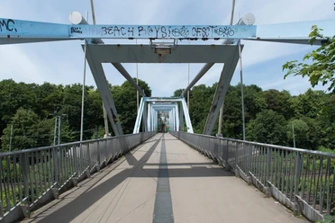 Heuschreckenbrücke Fußgängerbrücke mit stählernen Trägern und künstlerischem Graffiti, umgeben von grüner Landschaft.