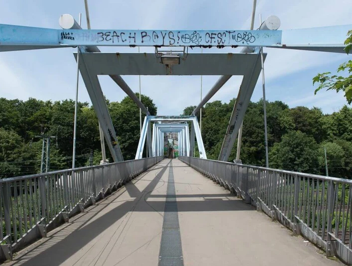 Heuschreckenbrücke Fußgängerbrücke mit stählernen Trägern und künstlerischem Graffiti, umgeben von grüner Landschaft.Pedestrian bridge with steel girders and artistic graffiti, surrounded by a green landscape.