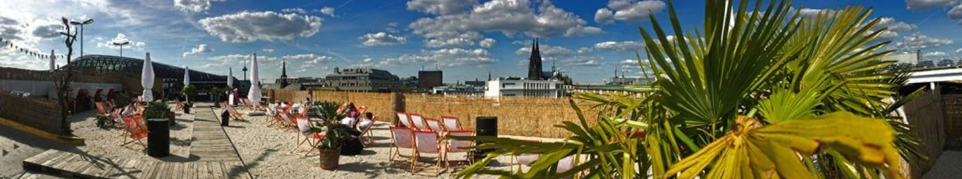 SonnenscheinEtage Auf dem Bild ist eine sonnige Dachterrasse zu sehen, die mit Sand bedeckt ist und Liegestühle bietet. Palmen stehen im Vordergrund, während sich im Hintergrund die Skyline von Köln mit seinem berühmten Dom abzeichnet. Fluffige Wolken ziehen am strahlend blauen Himmel vorüber.The picture shows a sunny roof terrace covered with sand and sun loungers. Palm trees stand in the foreground, while the skyline of Cologne with its famous cathedral looms in the background. Fluffy clouds pass by in the bright blue sky.