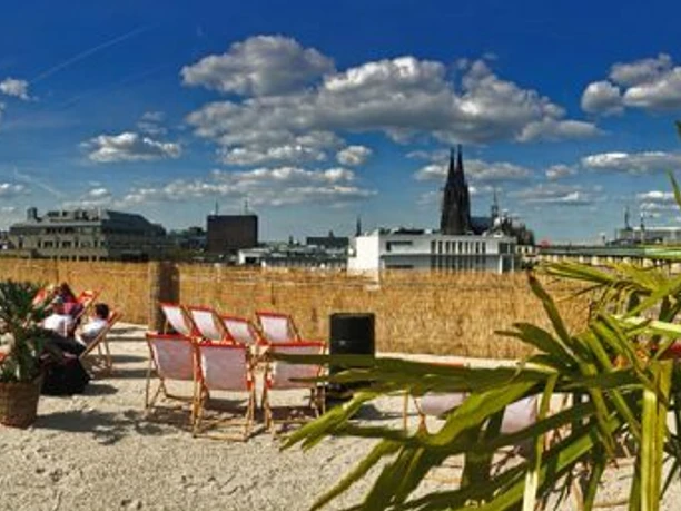 SonnenscheinEtage The picture shows a sunny roof terrace covered with sand and sun loungers. Palm trees stand in the foreground, while the skyline of Cologne with its famous cathedral looms in the background. Fluffy clouds pass by in the bright blue sky.
