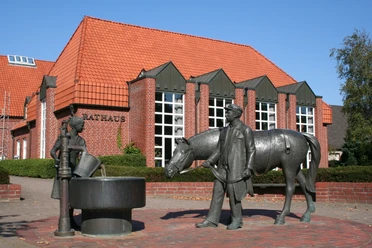 Gebouw van rode baksteen met een rood dak, ervoor een fontein met figuren van een paard en mensen.