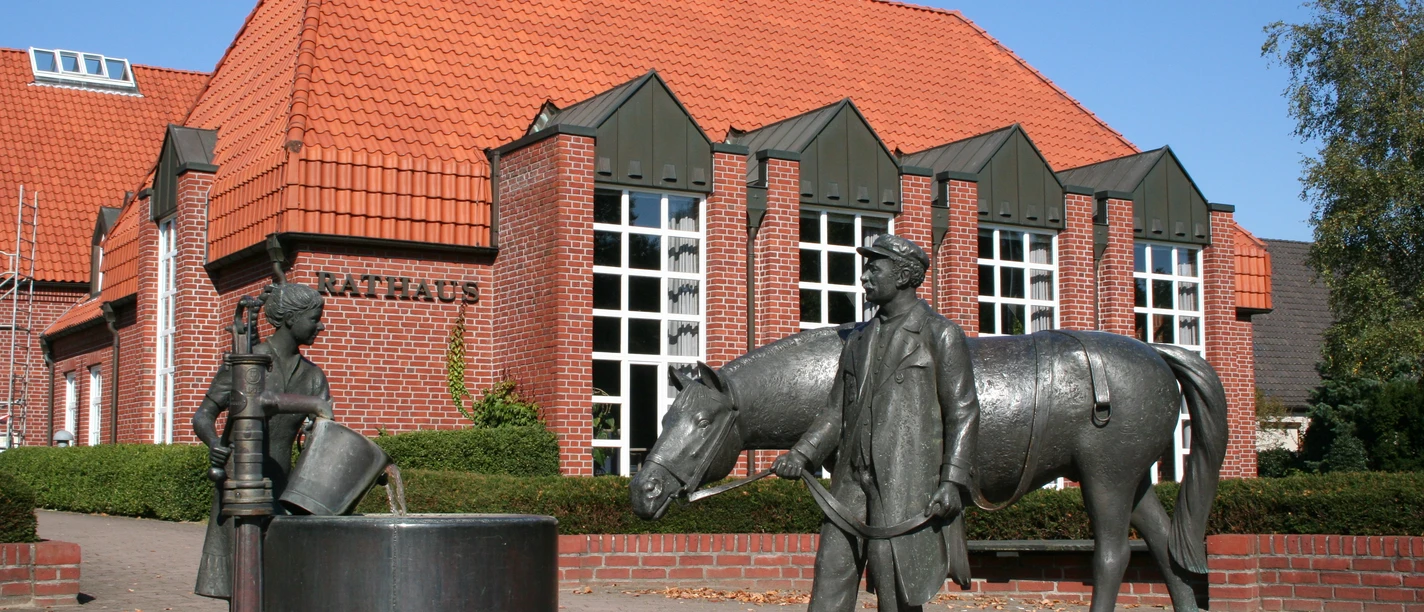 Gebouw van rode baksteen met een rood dak, ervoor een fontein met figuren van een paard en mensen.