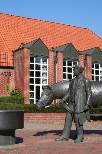 Rotes Backsteingebäude mit rotem Dach, davor Brunnen mit Figuren eines Pferdes und Menschen.