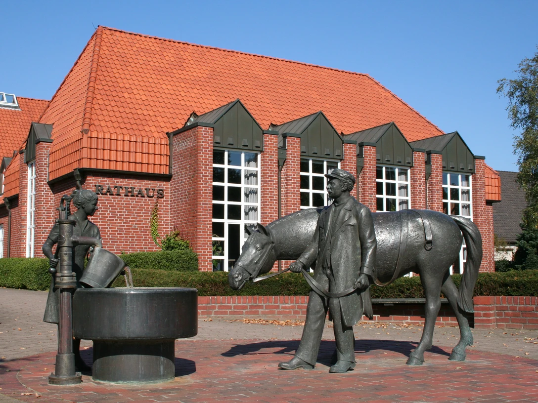 Rathaus Langwedel Rotes Backsteingebäude mit rotem Dach, davor Brunnen mit Figuren eines Pferdes und Menschen.