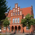 Rotes Backsteingebäude mit markantem Stufengiebel, umgeben von Bäumen und blauem Himmel.