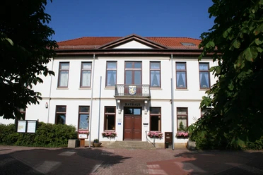 Das zweigeschossige, weiße Rathaus mit rotem Ziegeldach und zentralem Eingang ist von Bäumen umgeben.