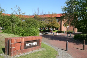 Backsteinbau des Rathauses Weyhe mit rotem Dach, umgeben von grünen Bäumen und blühenden Sträuchern.