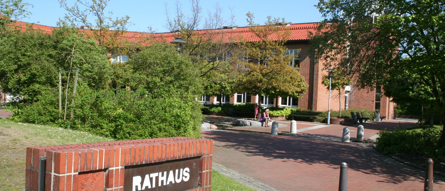 Rathaus Weyhe Backsteinbau des Rathauses Weyhe mit rotem Dach, umgeben von grünen Bäumen und blühenden Sträuchern.