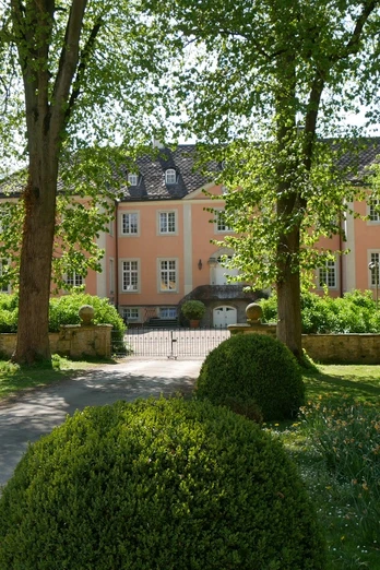 Helles Gebäude mit rosa Fassade, umgeben von gepflegtem Garten und großen, schattenspendenden Bäumen.