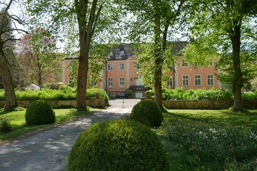 Schloss Rheder Helles Gebäude mit rosa Fassade, umgeben von gepflegtem Garten und großen, schattenspendenden Bäumen.