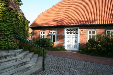 Ein traditionelles Backsteinhaus mit rotem Ziegeldach und weißen Fensterrahmen, umgeben von Pflanzen und Kopfsteinpflaster.