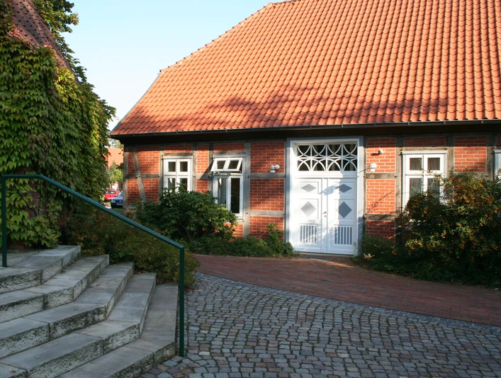 Ein traditionelles Backsteinhaus mit rotem Ziegeldach und weißen Fensterrahmen, umgeben von Pflanzen und Kopfsteinpflaster.