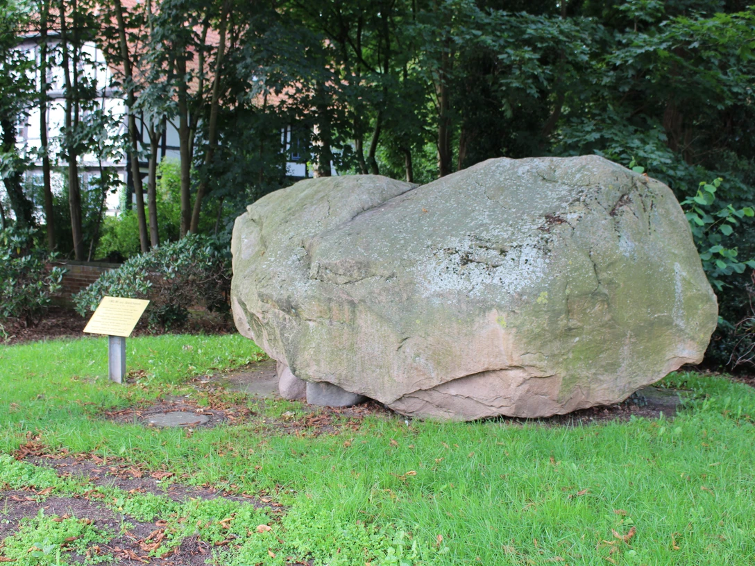 Ein großer Findling aus grauem Stein ruht auf grünem Gras, umgeben von Bäumen und Büschen.