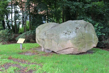 Findling Uchte Ein großer Findling aus grauem Stein ruht auf grünem Gras, umgeben von Bäumen und Büschen.