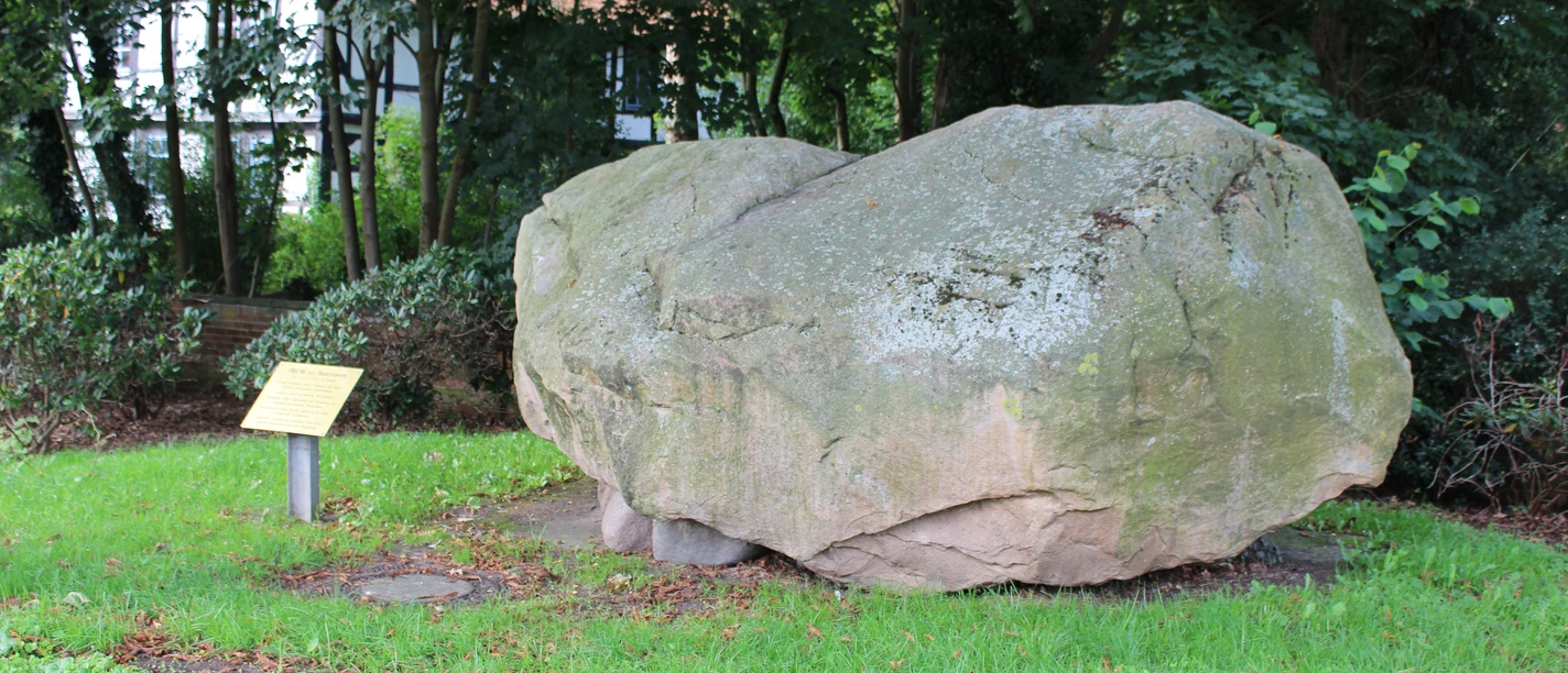 Findling Uchte Ein großer Findling aus grauem Stein ruht auf grünem Gras, umgeben von Bäumen und Büschen.