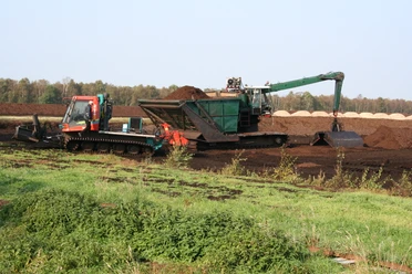 Moor Ein Torflader und ein Bagger arbeiten in einem weiten Moorgebiet unter klarem Himmel, umgeben von Vegetation.