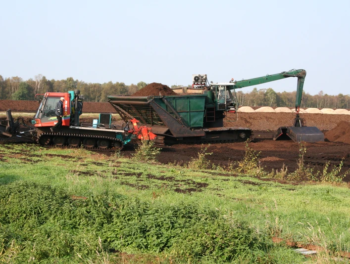 Moor Ein Torflader und ein Bagger arbeiten in einem weiten Moorgebiet unter klarem Himmel, umgeben von Vegetation.