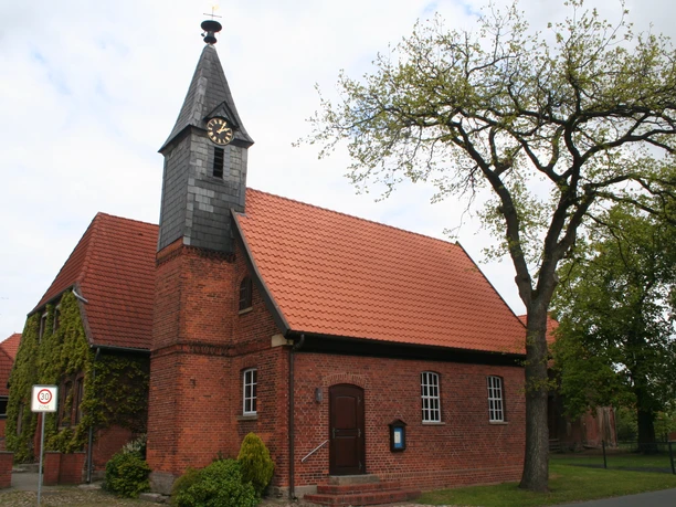 Rote Backsteinkapelle mit spitzem Turm und rotem Dach, von grünem Baum und bewölktem Himmel umgeben.