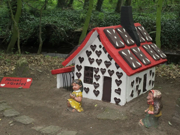 Das Bild zeigt ein kleines Lebkuchenhaus im Wald, verziert mit Herzformen, umgeben von Figuren.
