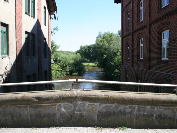 A view from a stone bridge of a river, framed by red brick buildings and trees.