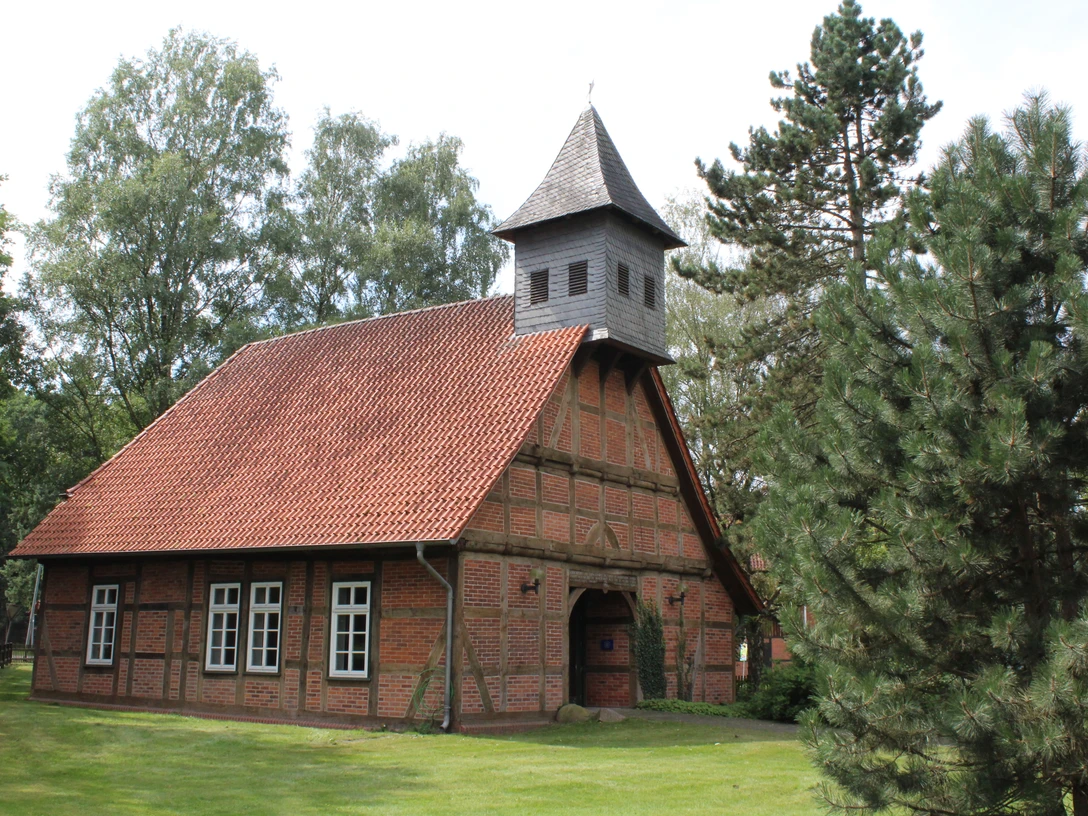 Backsteingebäude mit spitzem Turm und rotem Ziegeldach, umgeben von Bäumen und grüner Wiese.