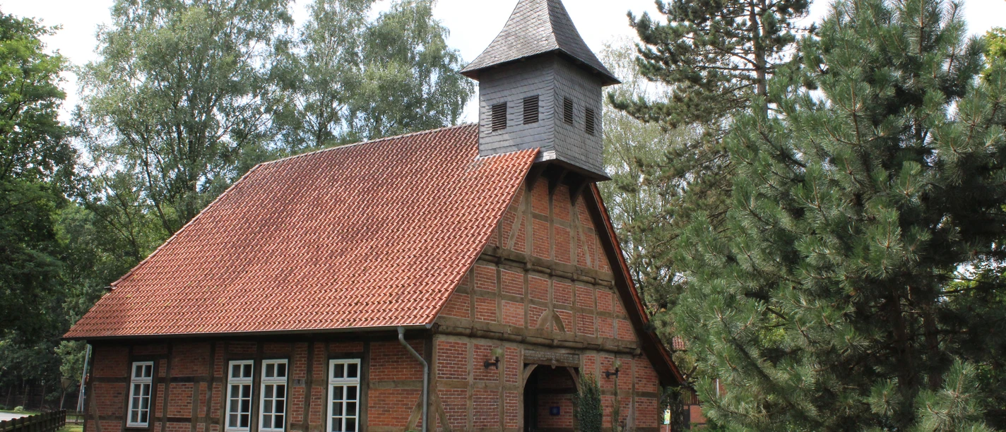 Backsteingebäude mit spitzem Turm und rotem Ziegeldach, umgeben von Bäumen und grüner Wiese.