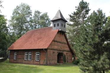 Alte Schule Sonnenborstel Backsteingebäude mit spitzem Turm und rotem Ziegeldach, umgeben von Bäumen und grüner Wiese.