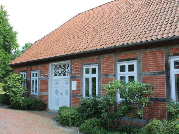Altes Küsterhaus Steimbke Backsteingebäude mit weißen Fensterrahmen und Ziegelwalmdach, umgeben von grünen Büschen.