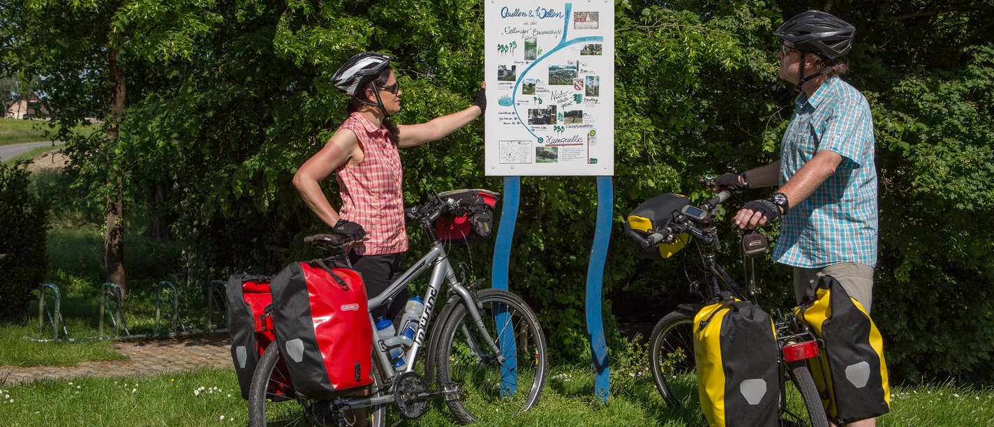 Radeln an der Bifurkation - Klaus Herzmann.JPG Zwei Radfahrer betrachten eine Infotafel an einem sonnigen Tag im Grünen.