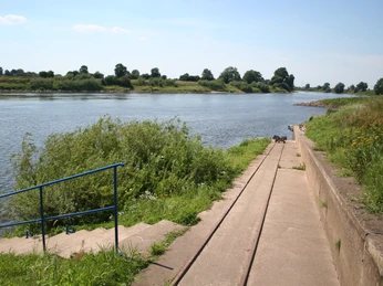 Eine Rampe mit Schienen führt zu einem Flussufer, umgeben von Grünflächen und Bäumen im Hintergrund.