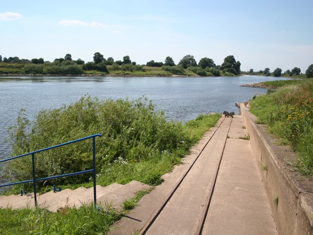 Intscheder Wehr Bootswagen Kanu Eine Rampe mit Schienen führt zu einem Flussufer, umgeben von Grünflächen und Bäumen im Hintergrund.