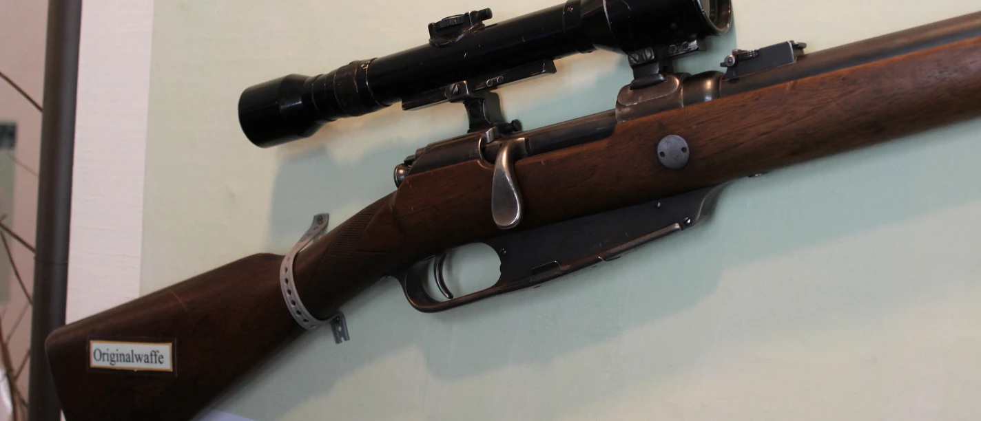 Gewehr Ein historisches Gewehr mit montiertem Zielfernrohr, ausgestellt vor einer erklärenden Tafel.