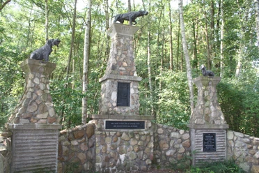 Hundedenkmal Drei steinerne Säulen mit bronzenen Hundeskulpturen stehen umgeben von dichtem Wald.