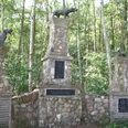 Hundedenkmal Drei steinerne Säulen mit bronzenen Hundeskulpturen stehen umgeben von dichtem Wald.
