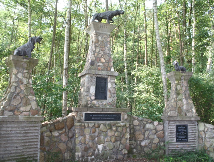 Hundedenkmal Drei steinerne Säulen mit bronzenen Hundeskulpturen stehen umgeben von dichtem Wald.