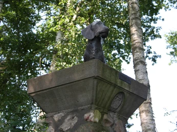 Hundedenkmal Hundeskulptur thront auf einem Steinsockel, umgeben von Bäumen im Hintergrund, sonniger Tag.