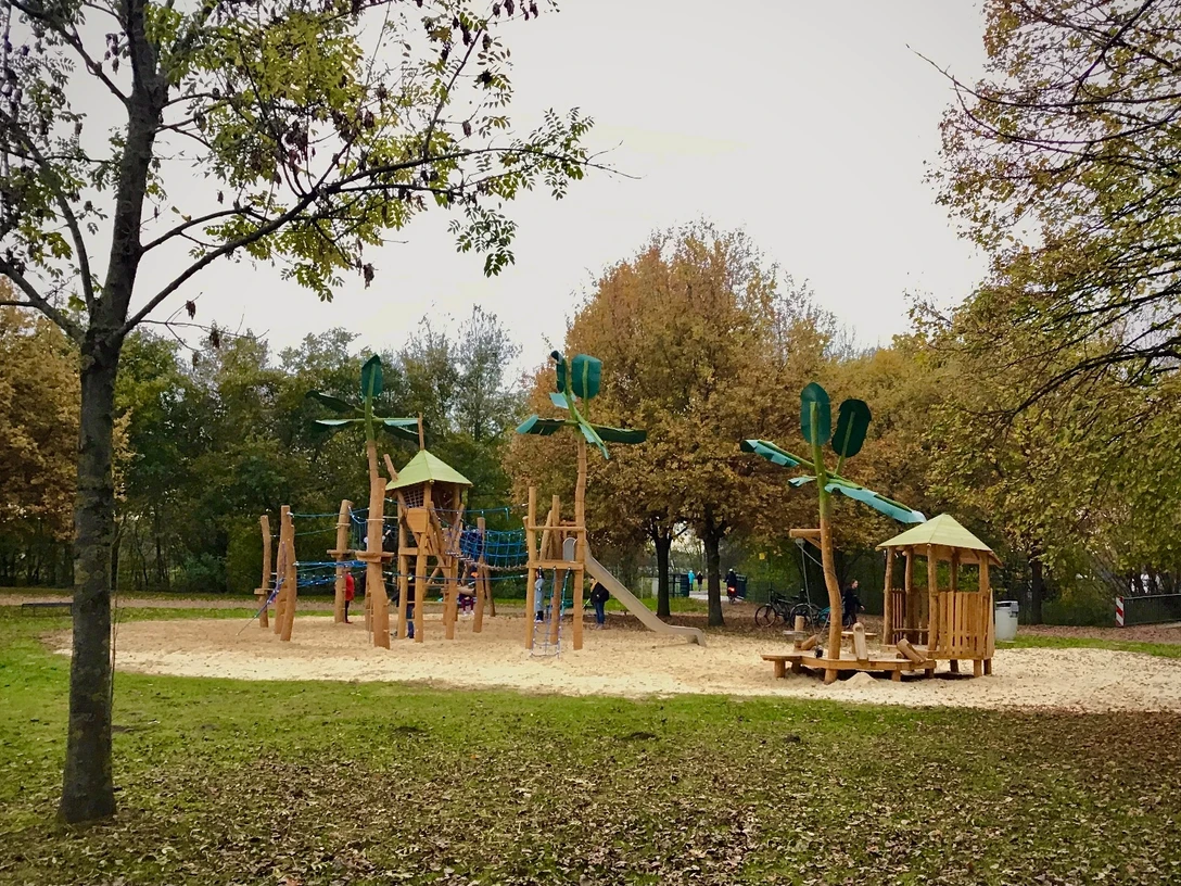 Holzspielplatz mit Klettergerüst und Rutschen, umgeben von Bäumen am Ufer gelegen.