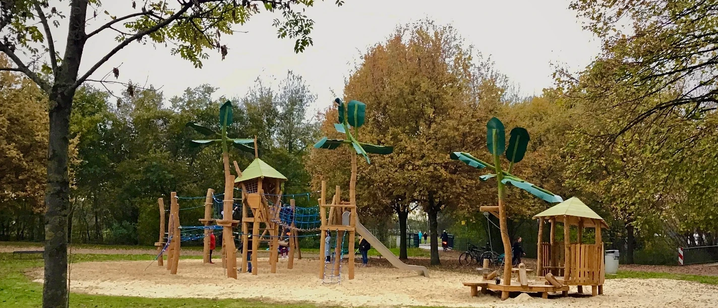 Holzspielplatz mit Klettergerüst und Rutschen, umgeben von Bäumen am Ufer gelegen.