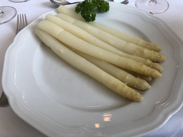 Fünf weiße Spargelstangen auf einem weißen Teller, dekoriert mit einem Zweig frischer Petersilie.