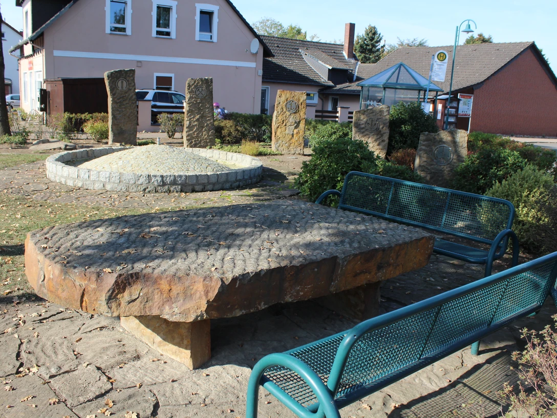 Brunnenplatz mit großen, flachen Natursteinen und modernen Bänken, umgeben von Sträuchern und Bäumen.