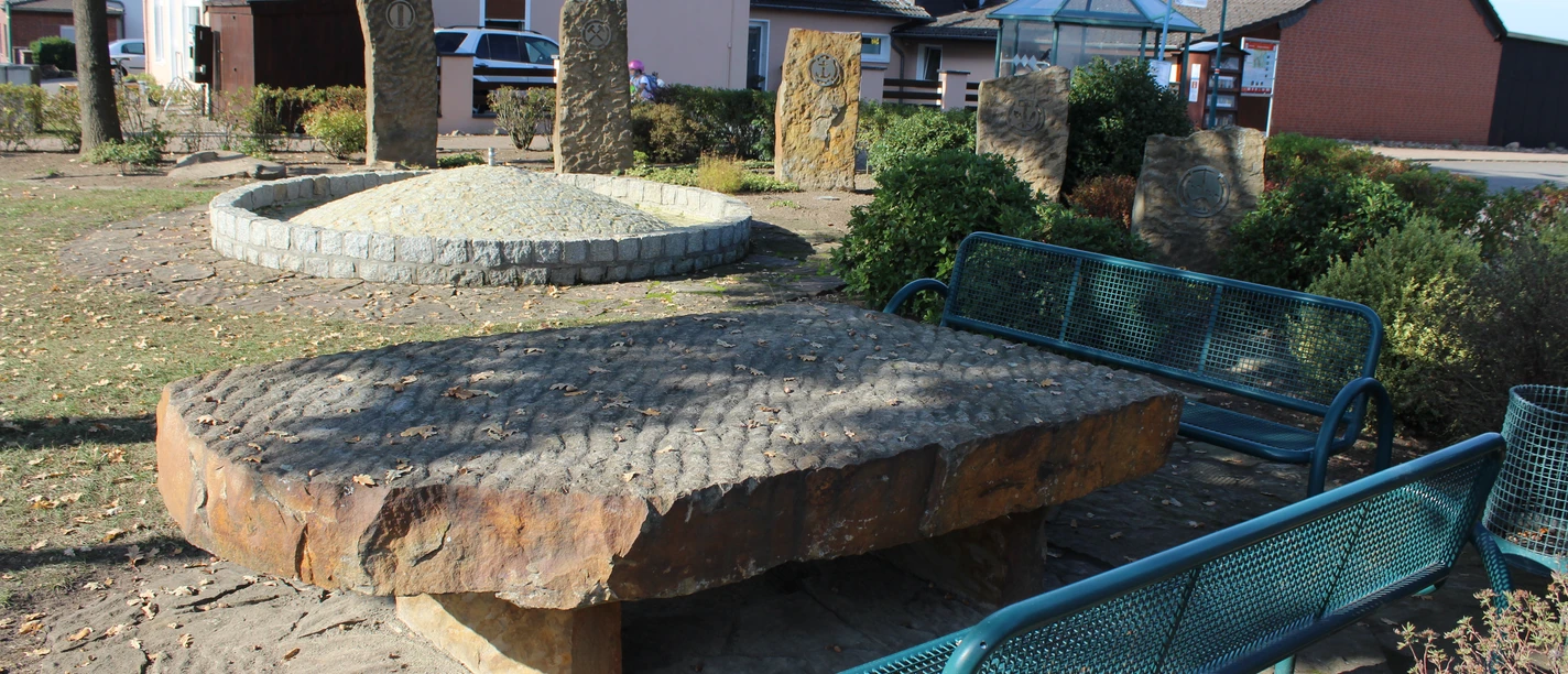 Brunnenplatz mit großen, flachen Natursteinen und modernen Bänken, umgeben von Sträuchern und Bäumen.