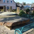Brunnen Münchehagen Brunnenplatz mit großen, flachen Natursteinen und modernen Bänken, umgeben von Sträuchern und Bäumen.
