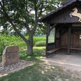 Holzhütte mit Wappenpferd, daneben ein Gedenkstein, umgeben von einem Kornfeld und grüner Natur.