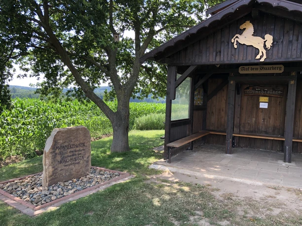 Holzhütte mit Wappenpferd, daneben ein Gedenkstein, umgeben von einem Kornfeld und grüner Natur.