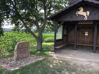Aussichtspunkt Haarberg Holzhütte mit Wappenpferd, daneben ein Gedenkstein, umgeben von einem Kornfeld und grüner Natur.
