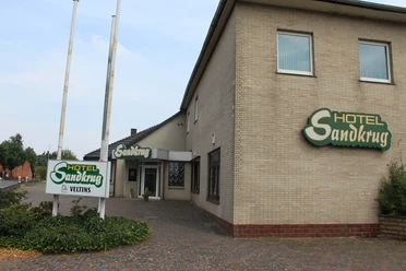 A large brick building of the Hotel Sandkrug with green signs and a cobblestone path.