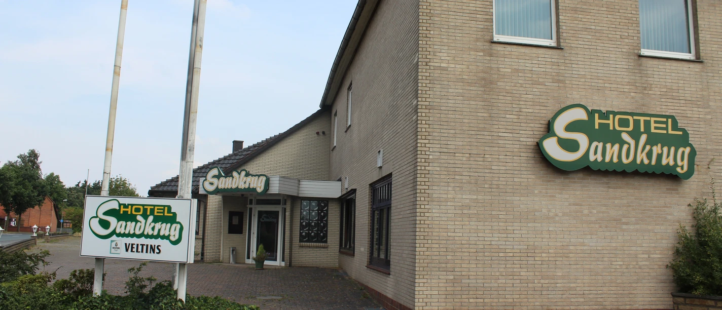 A large brick building of the Hotel Sandkrug with green signs and a cobblestone path.