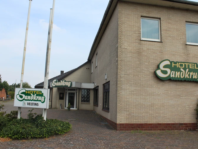 Ein großes Backsteingebäude des Hotel Sandkrug mit grünen Schildern und einem Kopfsteinpflasterweg.